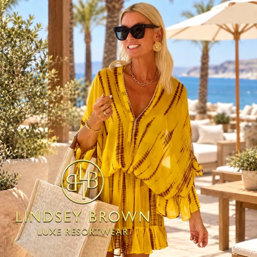 Lindsey Brown luxury resort wear branded designer silk and cotton kaftans collection image featuring a model wearing a vibrant yellow tie-dye silk kaftan dress with gold embellishments at a luxury beach resort.