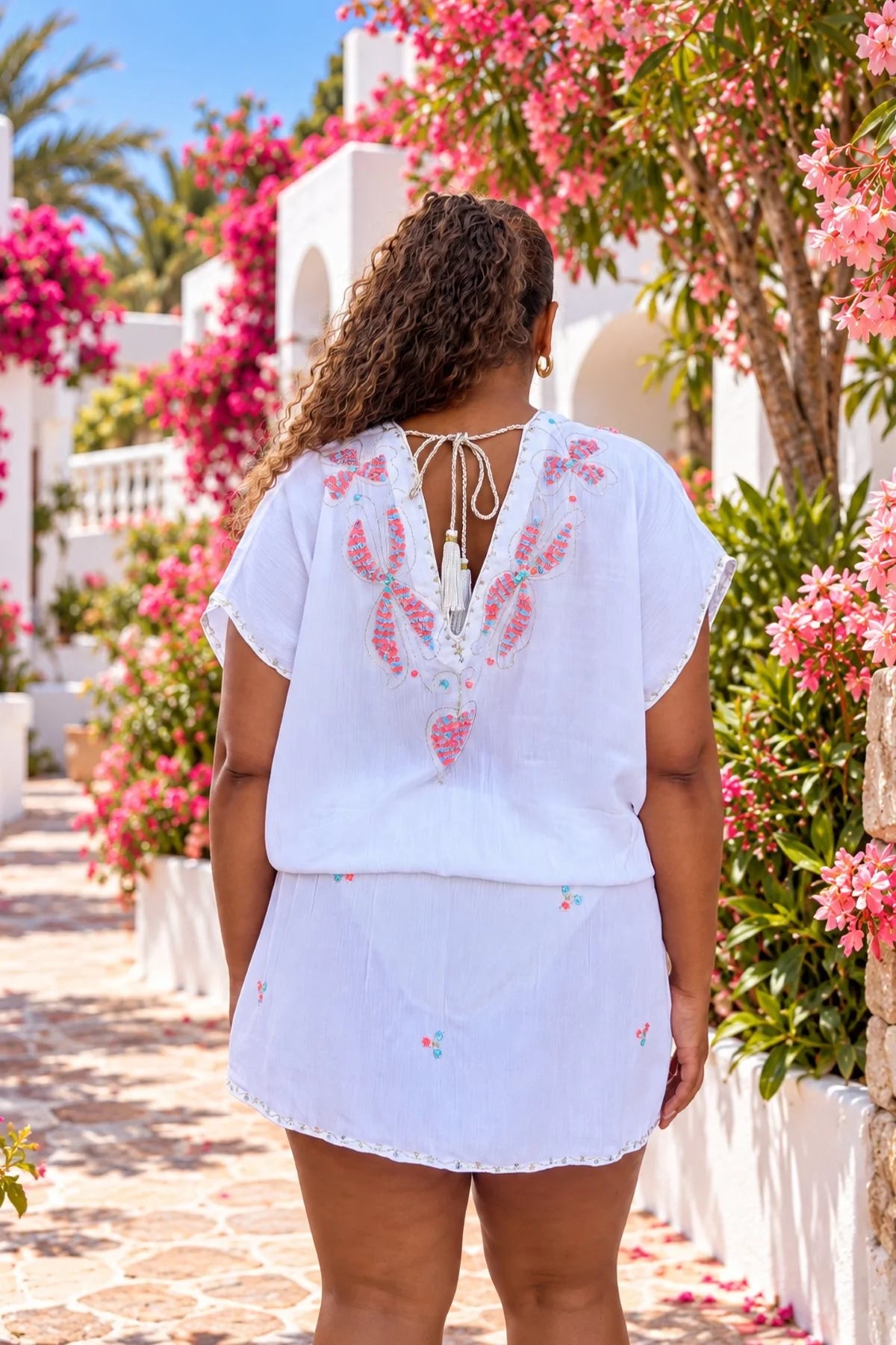 Lindsey Brown plus size luxury white cotton beach dress shown from the back, featuring delicate pink embroidery, a low V-back with tassel ties and a flattering elasticated waist.
