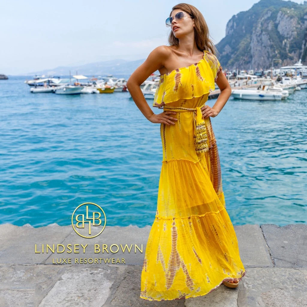 Model on a sunlit harborside wearing a Lindsey Brown luxury resort wear yellow tie-dye silk maxi resort dress with a one-shoulder neckline and belted waist, highlighting the luxury vacation style and bespoke styling concierge service offered by the fashion brand.
